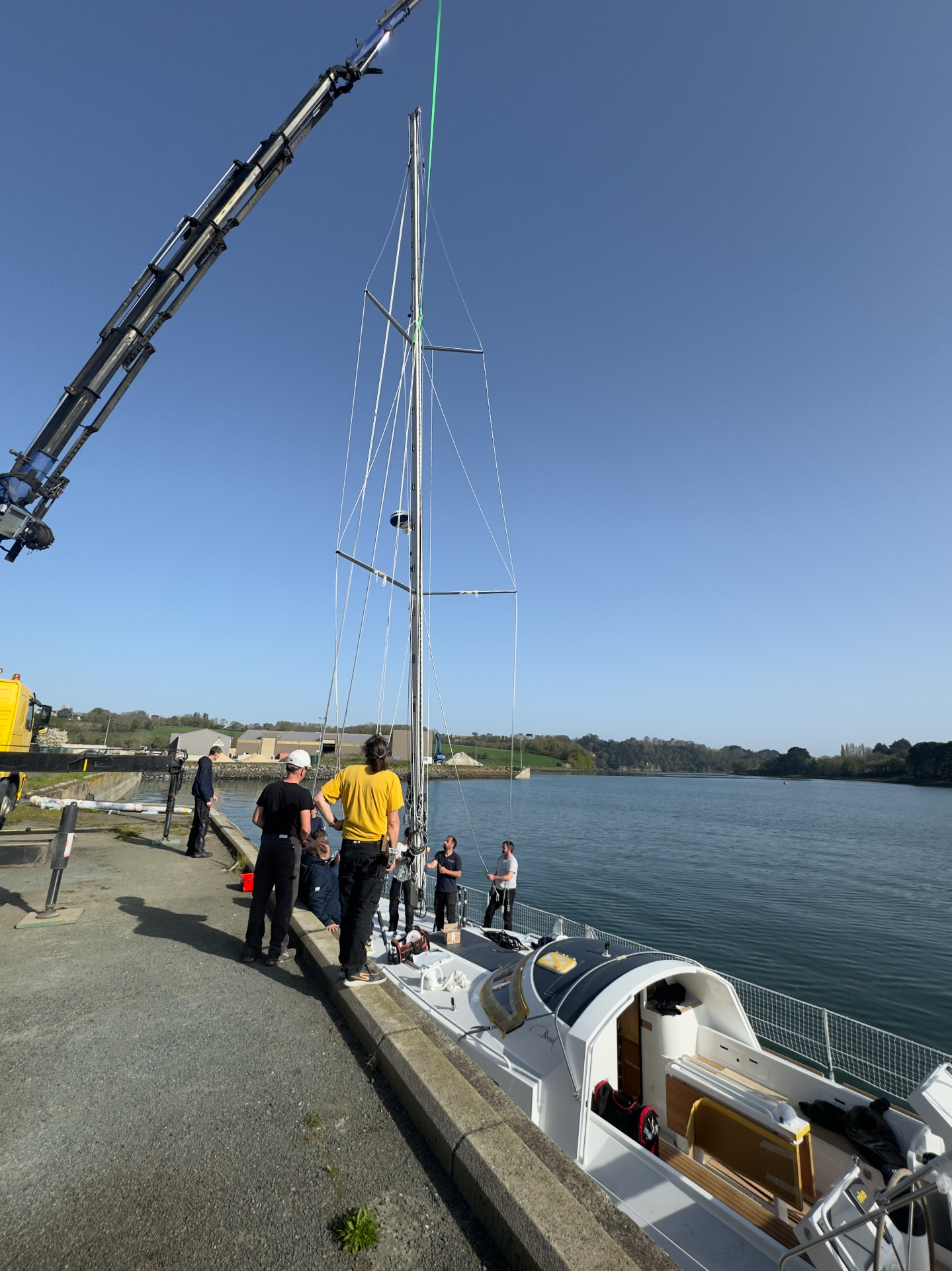 Crew members stand on a boat as a crane lowers a tall mast over calm river water.