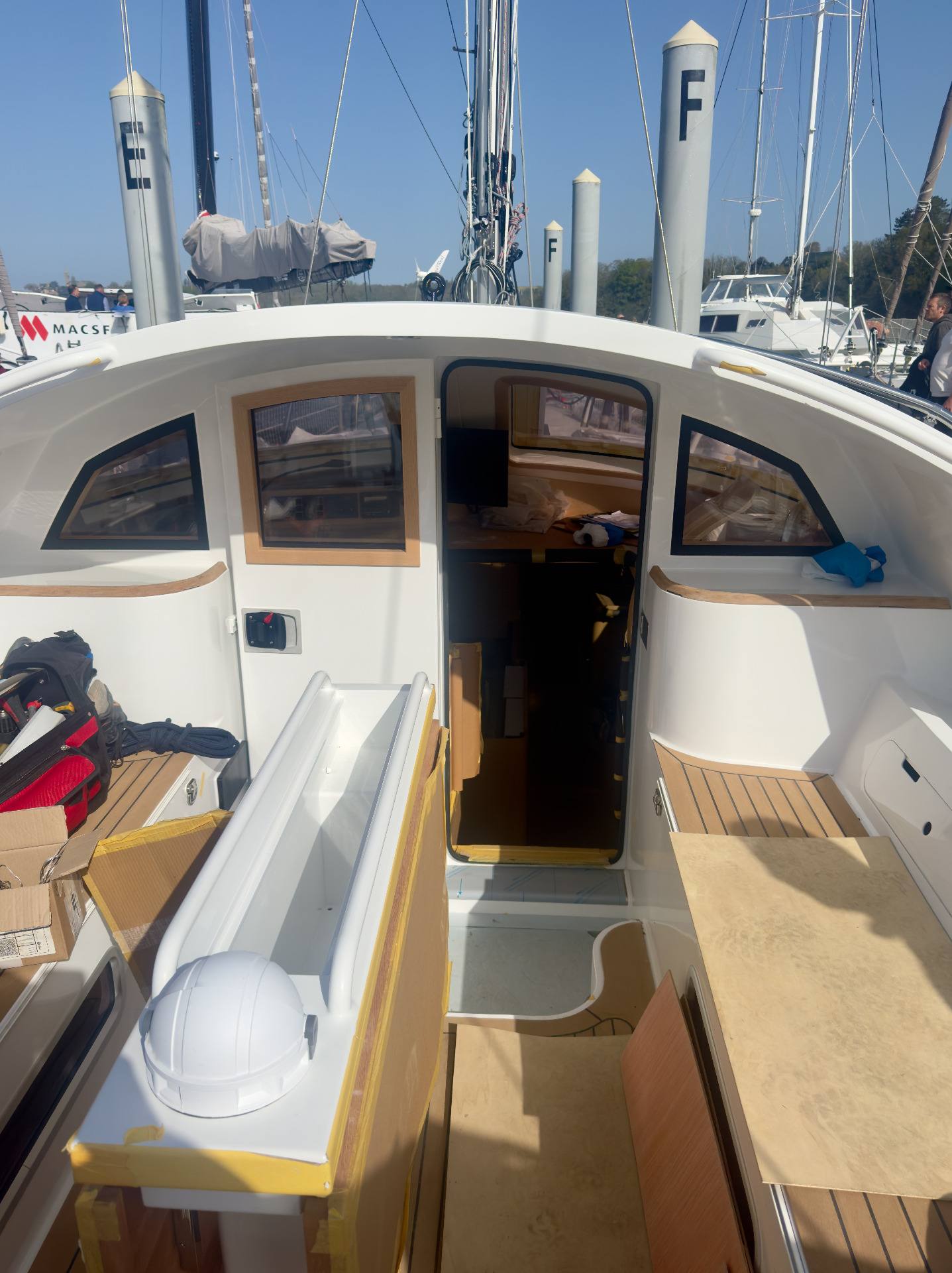 Open sailboat cockpit with wooden panels, gear inside, and tall masts on a sunny marina.